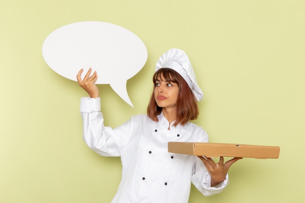 Photo gratuite vue avant femme cuisinier en costume de cuisinier blanc tenant la boîte de nourriture et panneau blanc sur un bureau vert