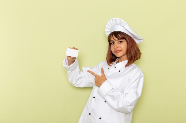 Vue avant femme cuisinier en costume de cuisinier blanc tenant une carte blanche sur une surface verte