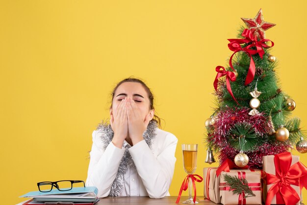 Vue avant femme médecin assis derrière la table avec des cadeaux de Noël et le bâillement d'arbre sur fond jaune