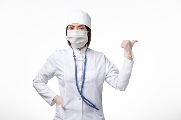 Vue avant femme médecin en costume médical blanc avec un masque en raison de la pandémie posant sur le mur blanc maladie médecine virus pandémie covid-