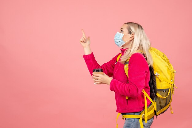 Vue avant femme voyageur avec sac à dos jaune portant un masque tenant une tasse de café doigt pointé vers le haut