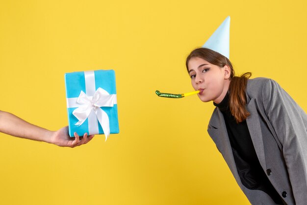 Vue avant heureuse jeune fille avec chapeau de fête à l'aide de bruiteur à la recherche de gifbox