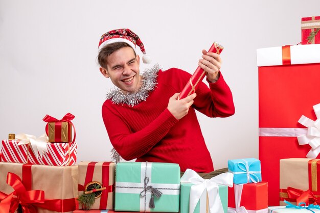 Vue avant heureux jeune homme à l'aide de popper de fête assis autour de cadeaux de Noël