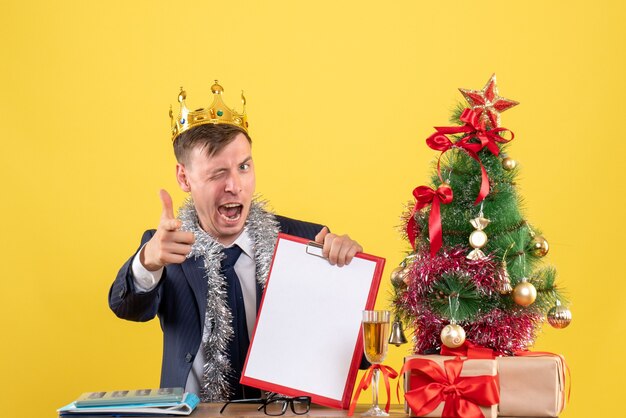 Vue avant de l'homme d'affaires clignote des yeux assis à la table près de l'arbre de Noël et présente sur fond jaune