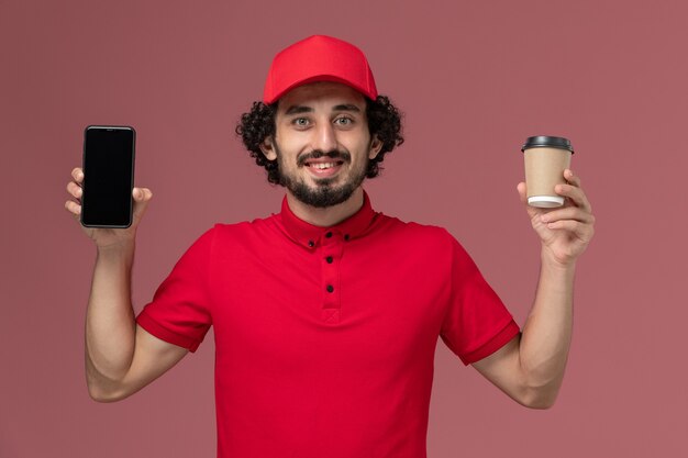 Vue avant de l'homme de livraison de messagerie en chemise rouge et cape tenant la tasse de café de livraison marron et téléphone sur mur rose clair employé de livraison de services