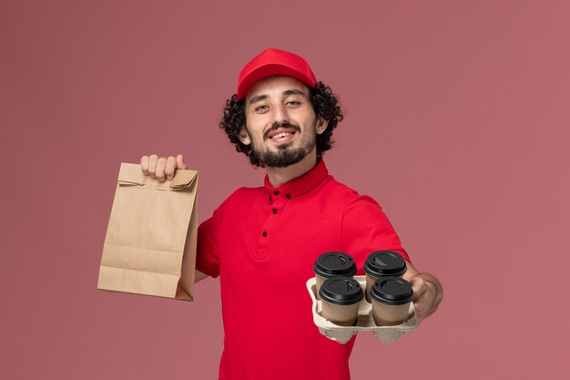 Vue avant de l'homme de livraison de messagerie en chemise rouge et cape tenant des tasses de café de livraison marron avec emballage alimentaire sur mur rose employé de livraison de service de travail