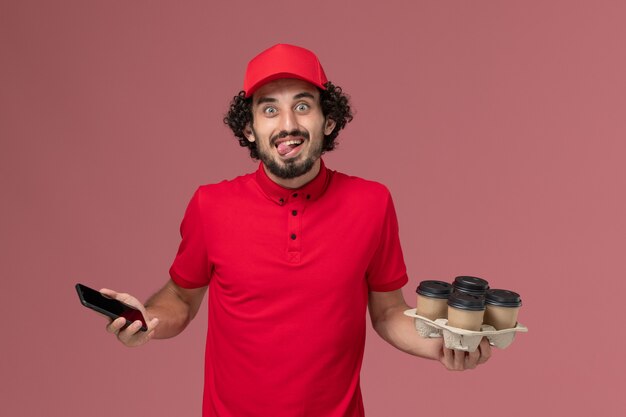 Vue avant de l'homme de livraison de messagerie en chemise rouge et cape tenant des tasses à café de livraison marron avec téléphone sur mur rose employé de livraison de services