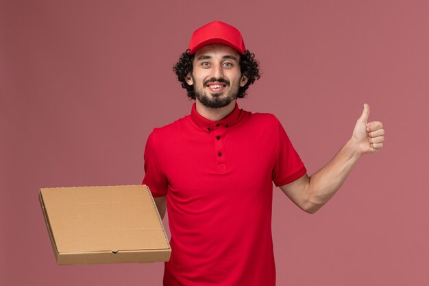 Vue avant de l'homme de livraison de messagerie homme en chemise rouge et cape tenant la boîte de nourriture de livraison sur le mur rose de l'entreprise de livraison de services employé travail mâle