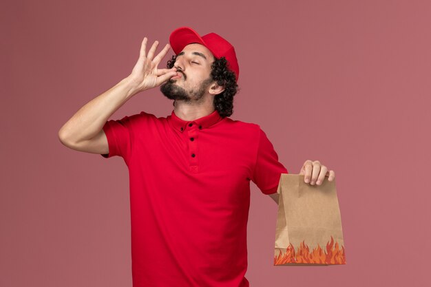 Vue avant de l'homme de livraison de messagerie homme en chemise rouge et cape tenant le paquet alimentaire sur le mur rose employé de l'entreprise de livraison de travailleur de service