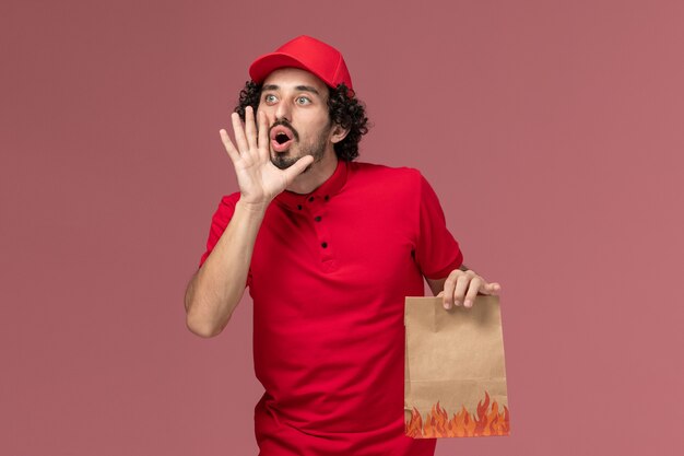 Vue avant de l'homme de livraison de messagerie homme en chemise rouge et cape tenant le paquet alimentaire papier et appelant quelqu'un sur l'employé de l'entreprise de livraison de services de bureau rose