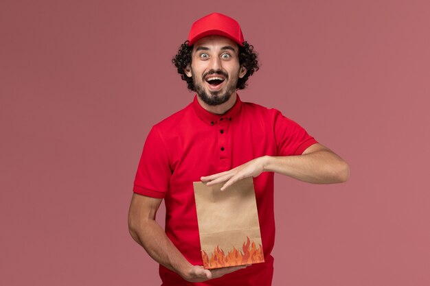 Vue avant de l'homme de livraison de messagerie homme en chemise rouge et cape tenant un paquet de papier alimentaire sur le mur rose de l'entreprise de livraison de services de travail des employés