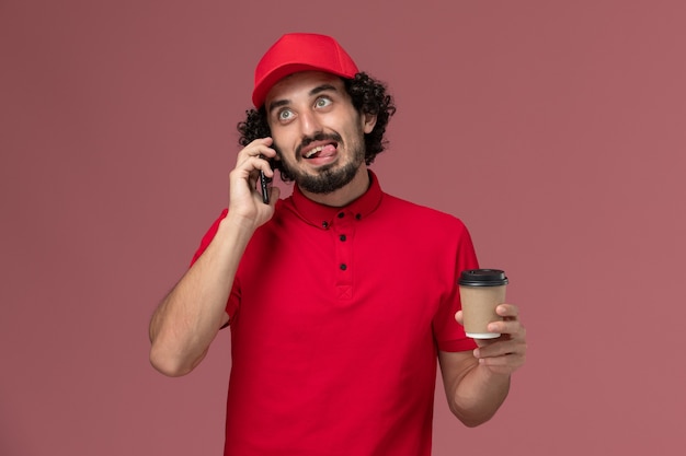 Vue avant de l'homme de livraison de messagerie homme en chemise rouge et cape tenant une tasse de café de livraison brune parler au téléphone sur le mur rose employé de livraison de services