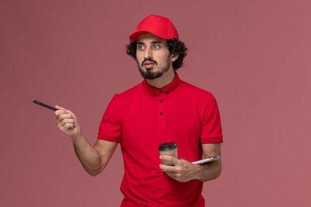 Vue avant de l'homme de livraison de messagerie homme en chemise rouge et cape tenant une tasse de café marron et bloc-notes avec stylo sur mur rose clair emploi employé de livraison de services