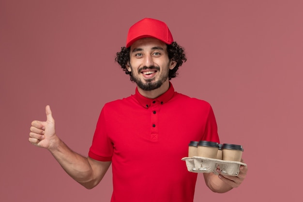 Vue avant de l'homme de livraison de messagerie homme en chemise rouge et cape tenant des tasses de café de livraison marron sur le mur rose clair employé de livraison de travail de service