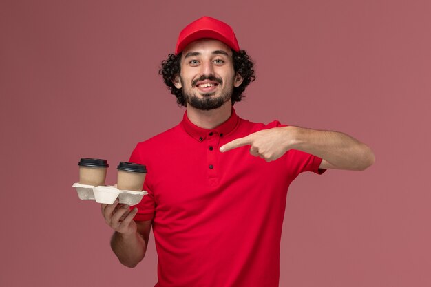 Vue avant de l'homme de livraison de messagerie homme en chemise rouge et cape tenant des tasses de café de livraison marron sur le mur rose employé de livraison de services