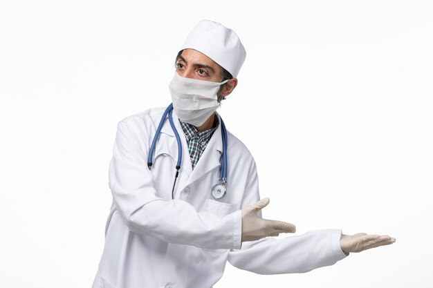 Vue avant de l'homme médecin en costume médical blanc et masque en raison d'un coronavirus sur un bureau blanc