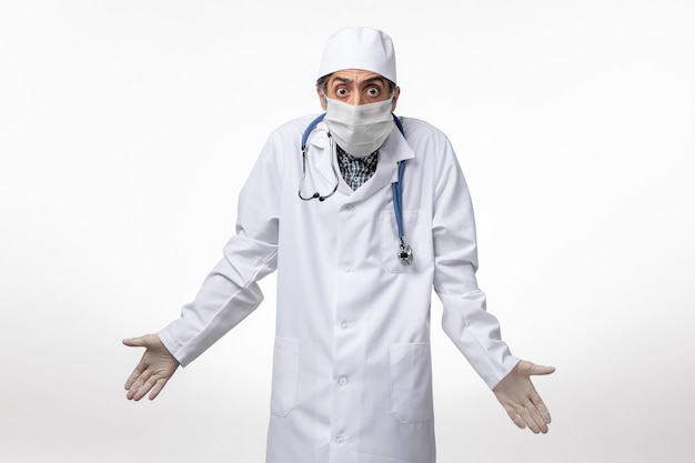 Vue avant de l'homme médecin en costume médical blanc avec masque en raison de coronavirus posant sur une surface blanche