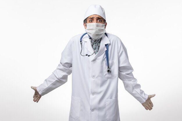 Vue avant de l'homme médecin en costume médical blanc avec masque en raison de coronavirus posant sur une surface blanche