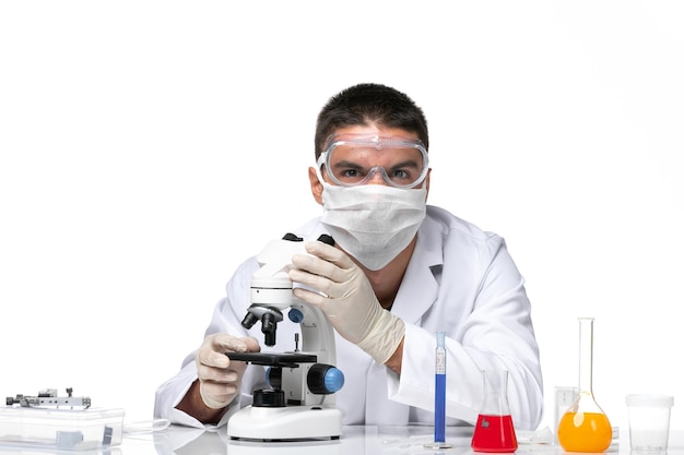 Vue avant de l'homme médecin en costume médical blanc avec masque en raison de covid à l'aide d'un microscope sur un espace blanc