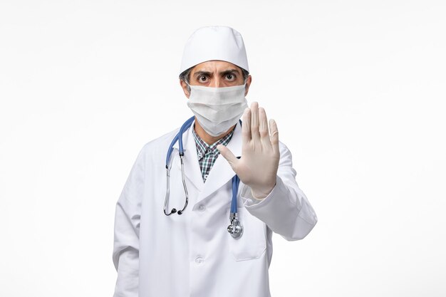 Vue avant de l'homme médecin en costume médical blanc avec masque en raison de covid montrant un panneau d'arrêt sur une surface blanche