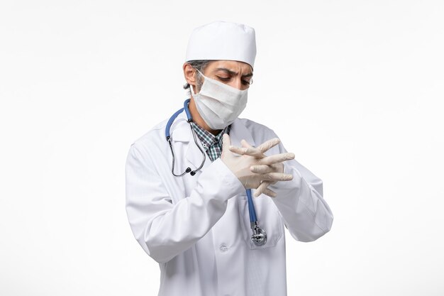 Vue avant de l'homme médecin en costume médical blanc avec masque en raison de covid portant des gants sur un bureau blanc
