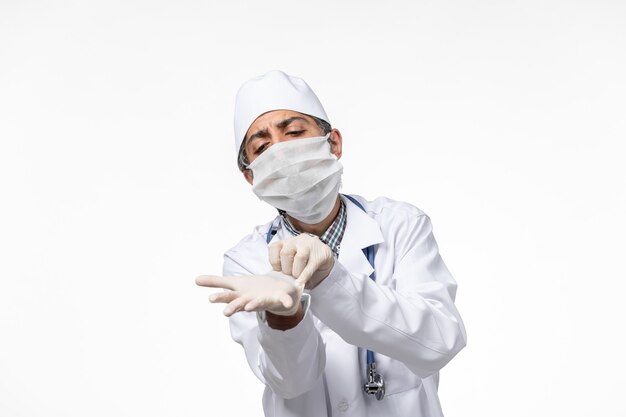 Vue avant de l'homme médecin en costume médical blanc avec masque en raison de covid portant des gants sur une surface blanche