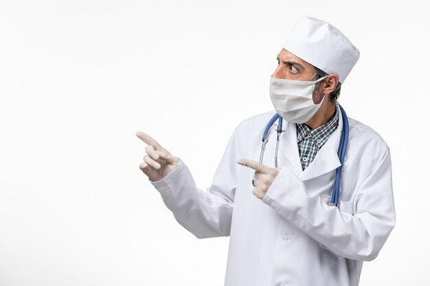 Vue avant de l'homme médecin en costume médical blanc avec masque en raison de covid sur une surface blanc clair