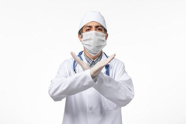 Vue avant de l'homme médecin en costume médical blanc avec masque en raison de covid sur une surface blanche