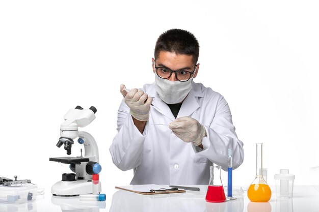 Vue avant de l'homme médecin en costume médical blanc avec masque en raison de covid travaillant avec des solutions sur un bureau blanc