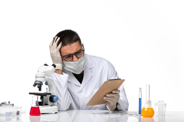 Vue avant de l'homme médecin en costume médical blanc avec masque en raison de l'écriture de notes covid sur l'espace blanc