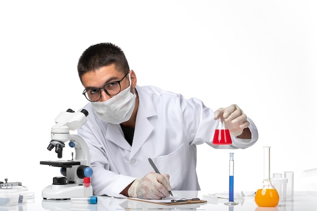 Vue avant de l'homme médecin en costume médical blanc et avec masque tenant une solution rouge sur un bureau blanc