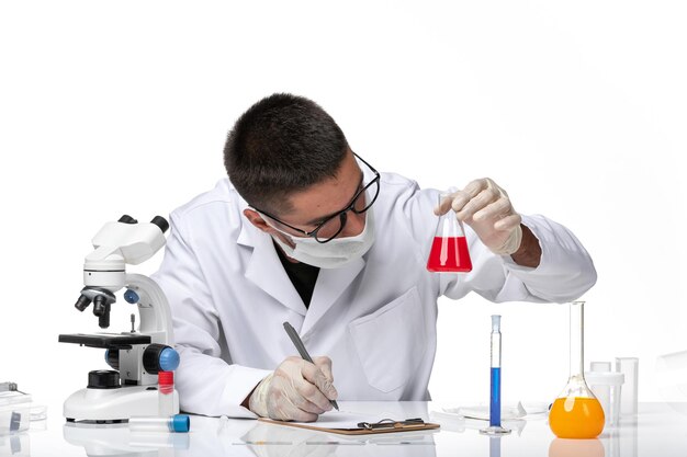 Vue avant de l'homme médecin en costume médical blanc et avec masque tenant une solution rouge sur un espace blanc