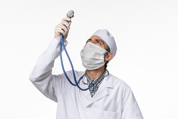 Vue avant de l'homme médecin en costume médical blanc portant un masque en raison de coronavirus tenant un stéthoscope sur une surface blanche