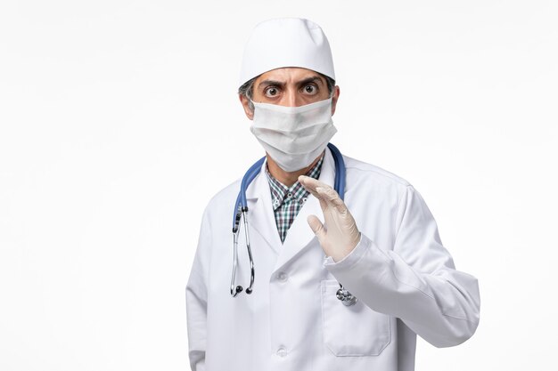 Vue avant de l'homme médecin en costume médical blanc portant un masque en raison de covid sur un bureau blanc clair