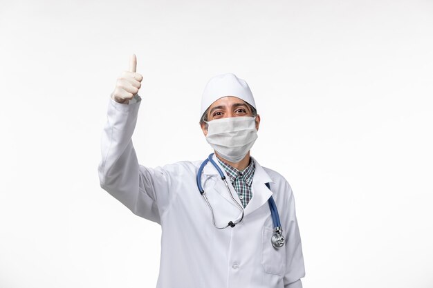 Vue avant de l'homme médecin en costume médical blanc portant un masque en raison de covid- souriant sur une surface blanche