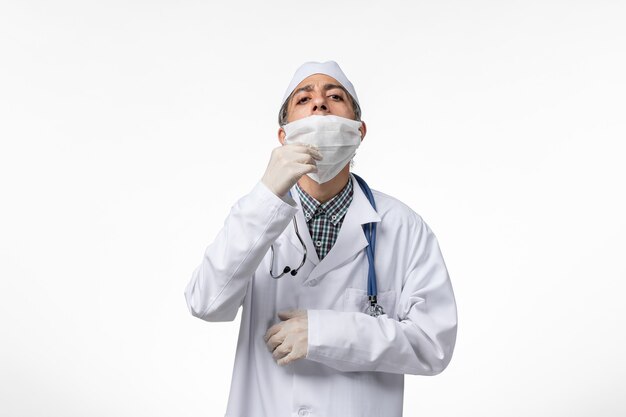 Vue avant de l'homme médecin en costume médical blanc en raison d'un coronavirus sur une surface blanche