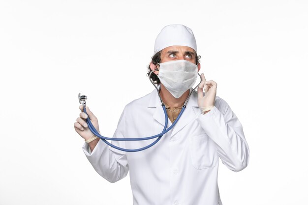 Vue avant de l'homme médecin en costume médical avec masque comme protection contre covid- à l'aide d'un stéthoscope sur la pandémie de coronavirus splash virus mur blanc