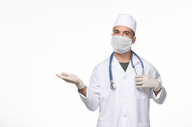 Vue avant de l'homme médecin en costume médical et masque contre le coronavirus à l'aide d'un stéthoscope sur un bureau blanc pandémie de maladie de maladie à virus covid-