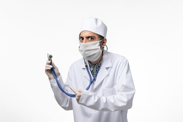 Vue avant de l'homme médecin en costume médical et masque en raison d'un coronavirus à l'aide d'un stéthoscope sur un bureau blanc