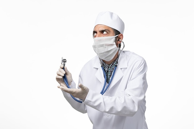 Vue Avant De L'homme Médecin En Costume Médical Et Masque En Raison D'un Coronavirus à L'aide D'un Stéthoscope Sur La Surface Blanche Claire