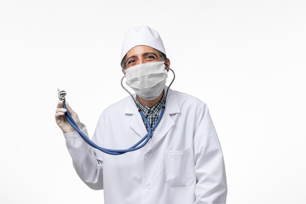Vue avant de l'homme médecin en costume médical et masque en raison d'un coronavirus à l'aide d'un stéthoscope sur une surface blanche