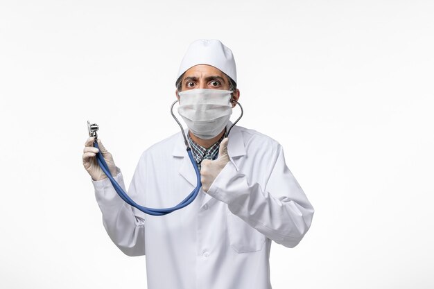 Vue avant de l'homme médecin en costume médical et masque en raison d'un coronavirus à l'aide d'un stéthoscope sur une surface blanche