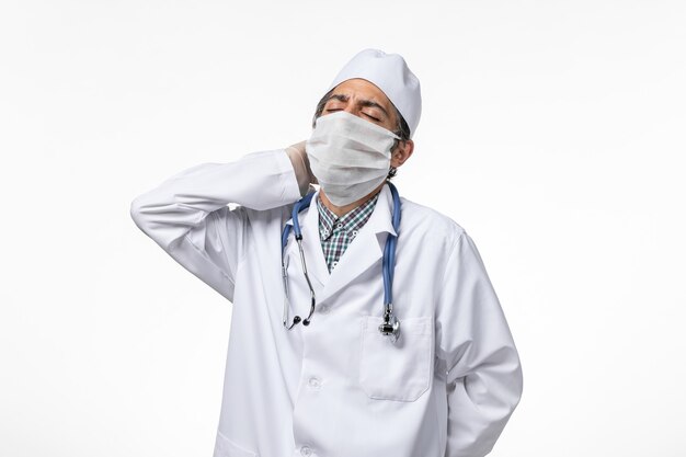 Vue avant de l'homme médecin en costume médical et masque en raison de coronavirus souffrant de maux de cou sur une surface blanche