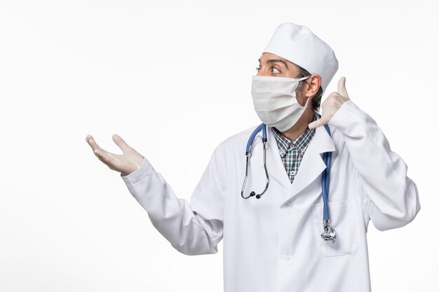 Vue avant de l'homme médecin en costume médical avec masque en raison de covid- posant sur une surface blanche