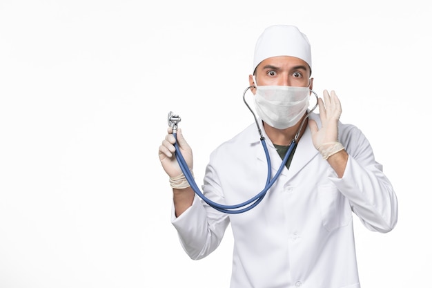 Vue avant de l'homme médecin en costume médical et portant un masque en raison de covid- à l'aide d'un stéthoscope sur mur blanc covid- maladie pandémique