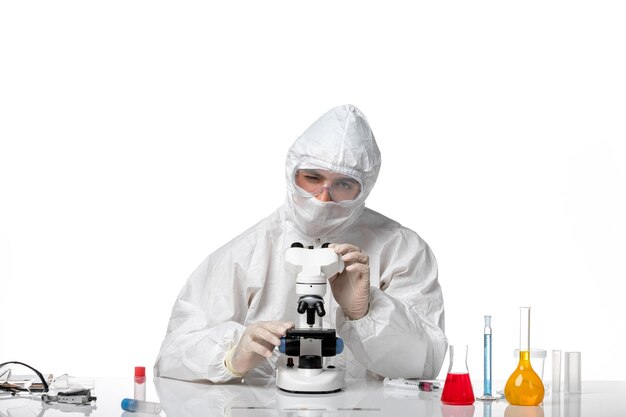 Vue avant de l'homme médecin en tenue de protection et avec masque à l'aide d'un microscope sur un bureau blanc