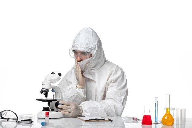 Vue avant de l'homme médecin en tenue de protection et avec masque à l'aide d'un microscope sur un bureau blanc