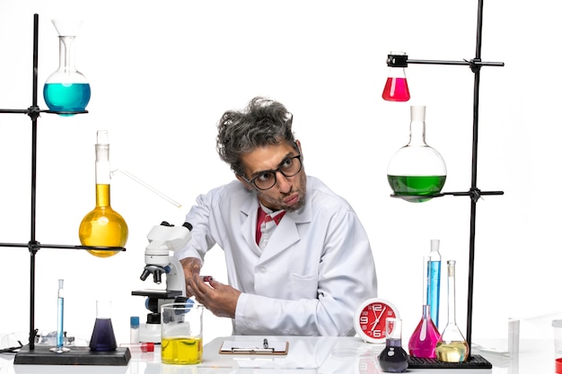 Vue avant de l'homme scientifique en costume médical assis en face de la table avec des solutions sur le fond blanc clair covid- laboratoire chimie de la santé des virus