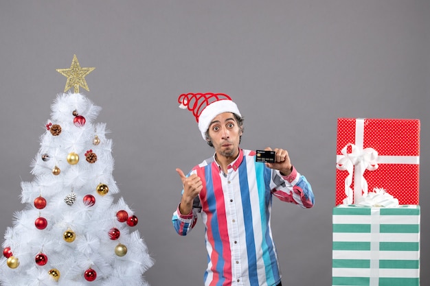 Vue avant de l'homme surpris avec carte de crédit montrant l'arbre de Noël debout près de différents cadeaux