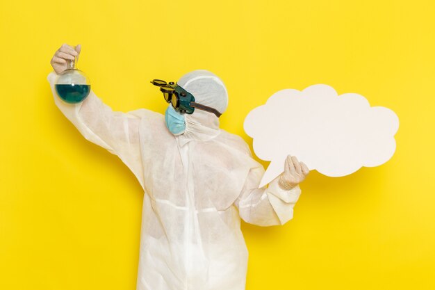 Vue avant de l'homme travailleur scientifique en costume spécial holding flask avec une solution verte et grand panneau blanc sur la surface jaune